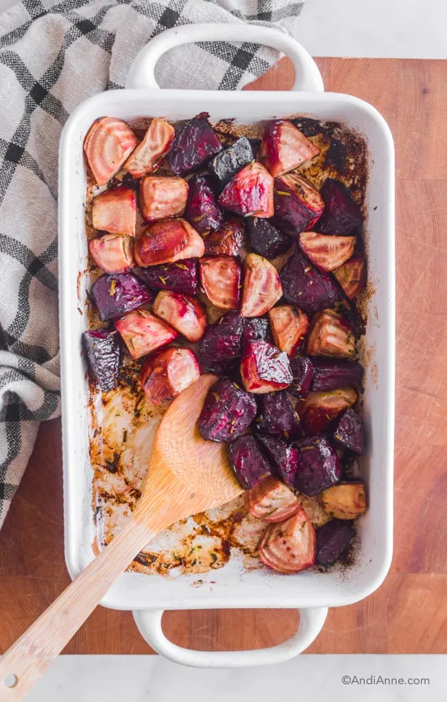 Roasted beets in a white casserole dish with a wooden spoon. A kitchen towel is beside it.