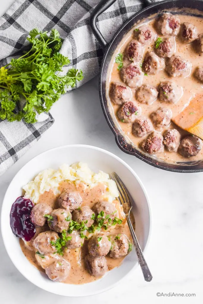 White dish with swedish meatballs and mashed potatoes with a fork. Black skillet beside with leftover meatballs.