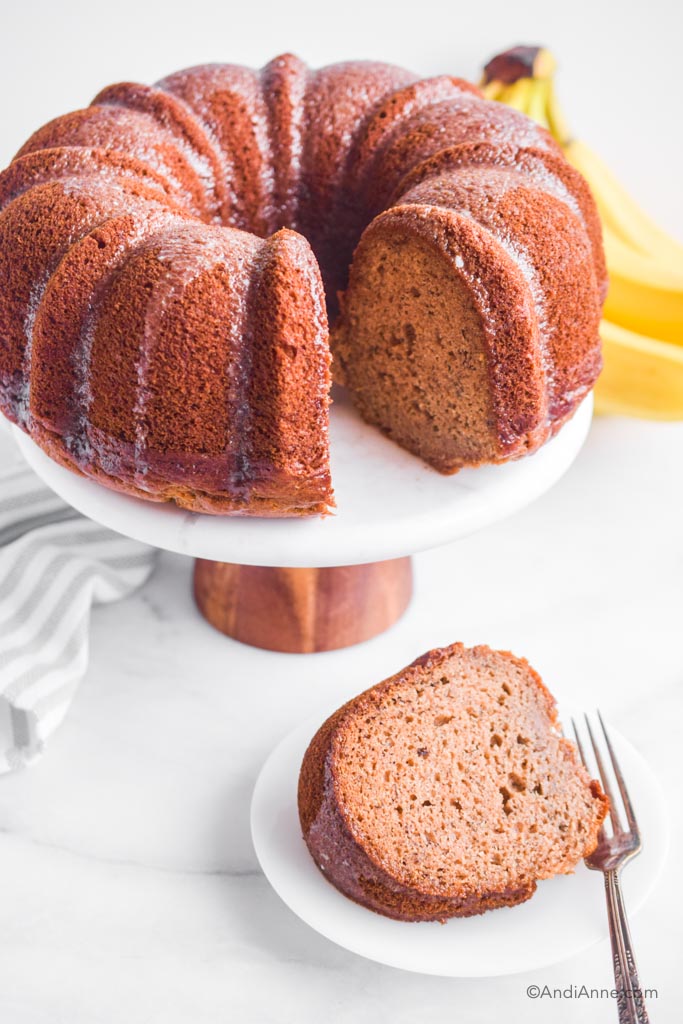 banana bundt cake on cake stand with bananas in background and slice of cake on plate with a fork.