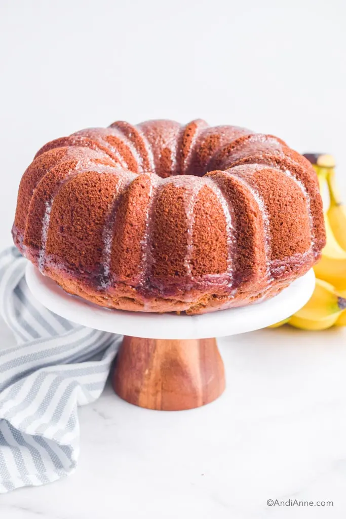 Banana bundt cake on cake stand with glaze drizzled overtop. Bananas and kitchen towel in background.
