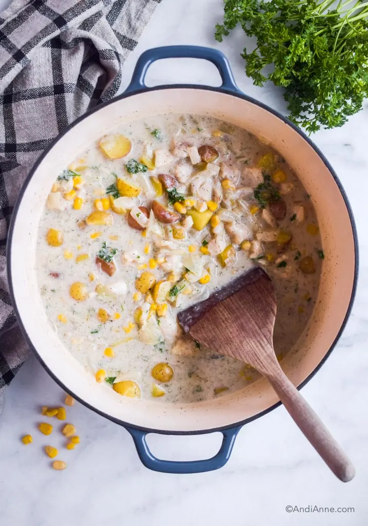 cod potato chowder in a dutch oven pot with wood spoon. Kitchen towel and parsley surround it.