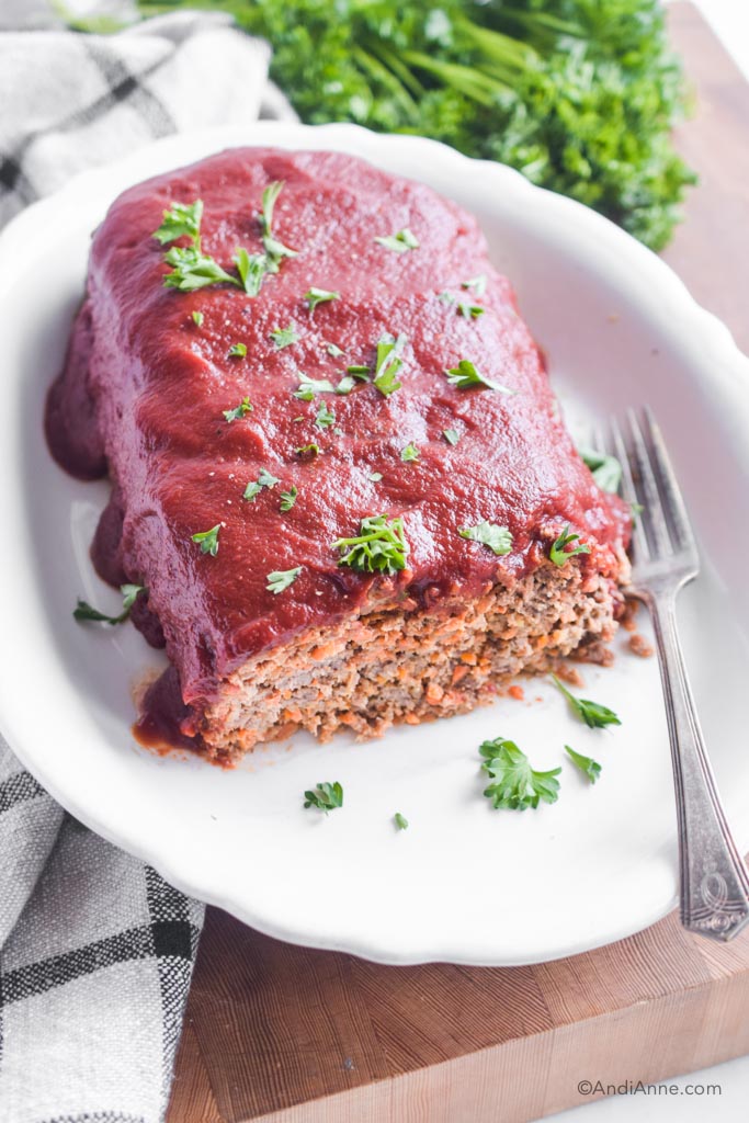Meatloaf covered in tomato sauce in a white dish with a fork and parsley sprinkled overtop.