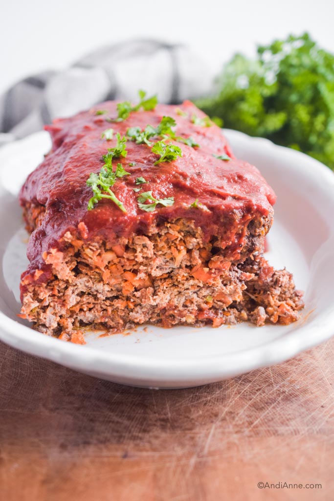 keto vegetable meatloaf with sprinkled parsley on top sitting in a white dish with a kitchen towel blurred in background.