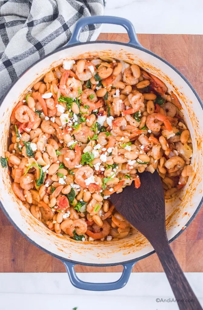 Looking down at one pot shrimp and bean dinner recipe with a wooden spatula. All in blue and white cast iron pot.