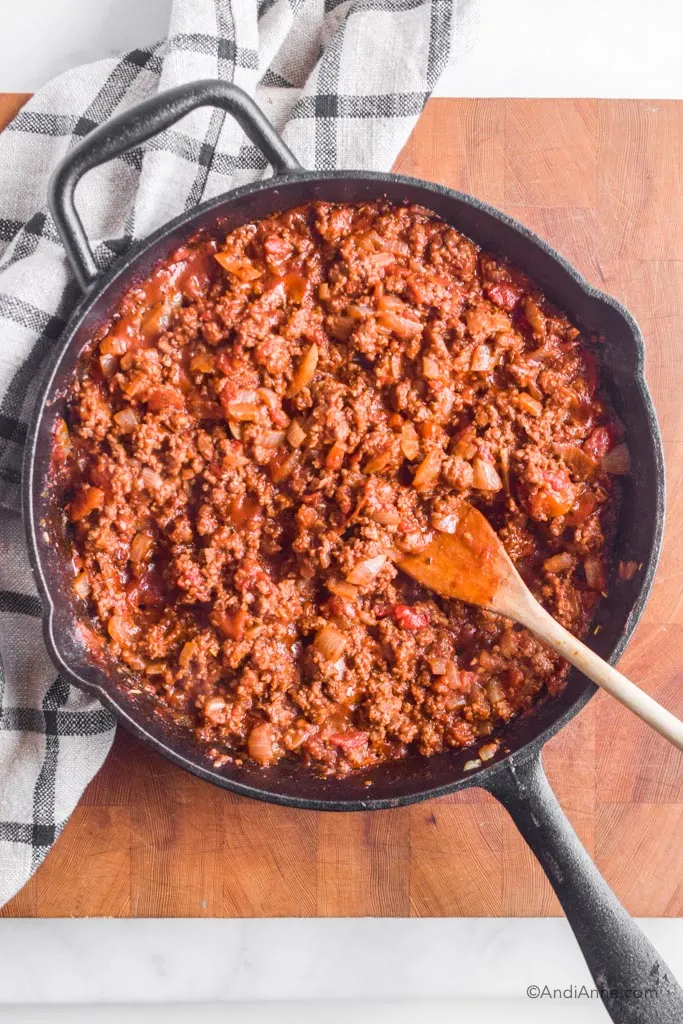 Cast iron skillet with bolognese sauce and a wood spatula. Pan is on a butcher block with kitchen towel beside it.