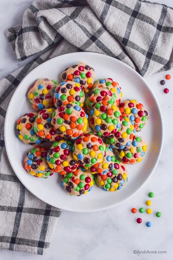 Cookies loaded with mini M&Ms on a white plate with kitchen towel and a few mini M&Ms beside plate.