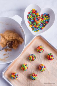 white bowl of batter with cookie scoop. Heart shaped bowl with mini M&Ms, cookie sheet with balls of cookie dough covered in M&Ms
