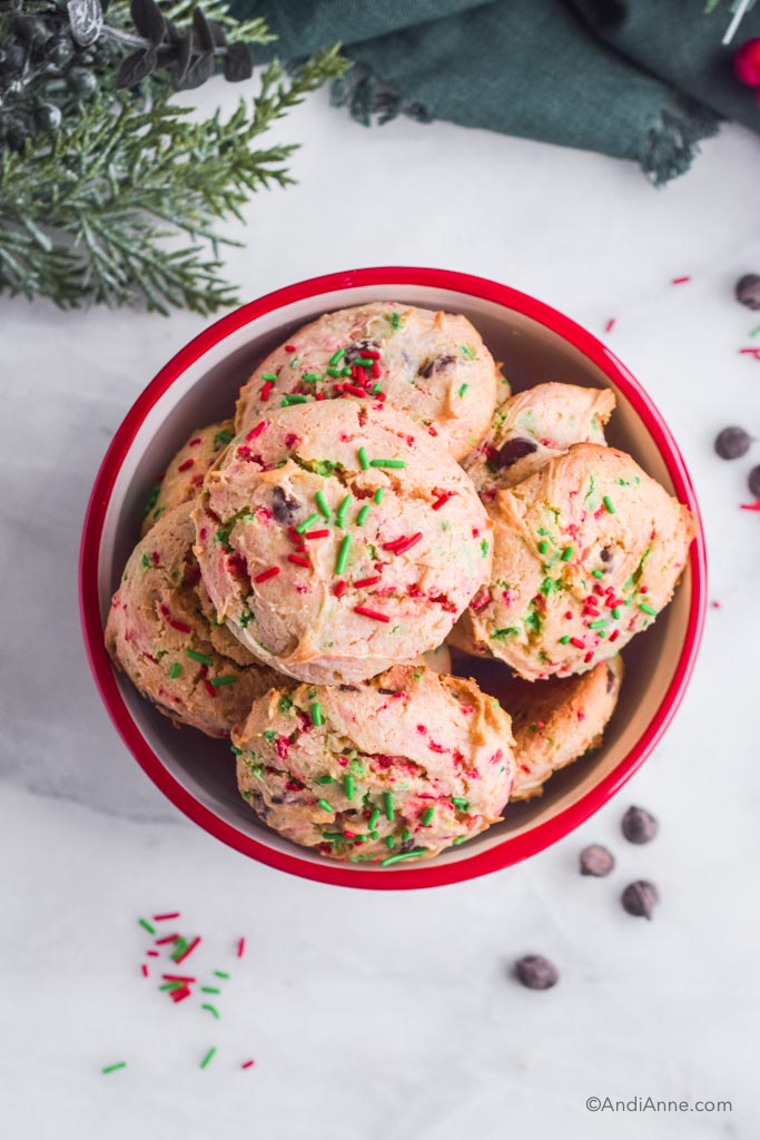 Pile of red and green santa cookies inside a red and white bowl. Chocolate chips and sprinkles outside of bowl. 
