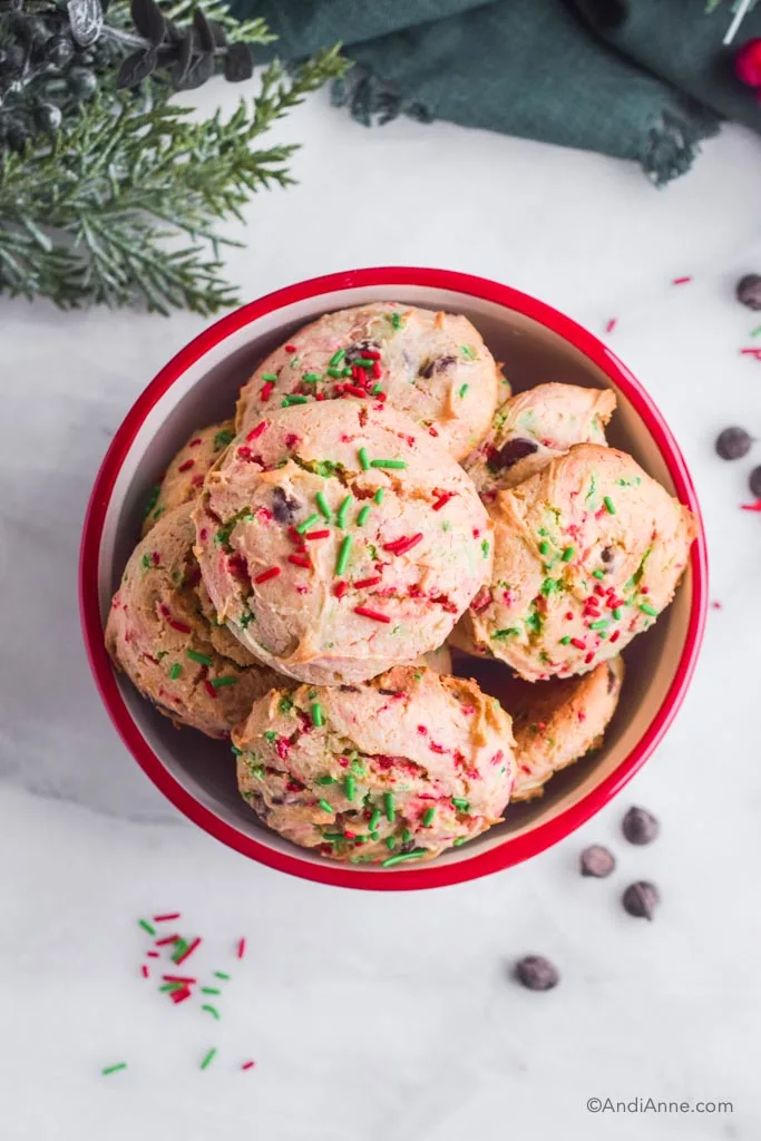Pile of red and green santa cookies inside a red and white bowl. Chocolate chips and sprinkles outside of bowl.