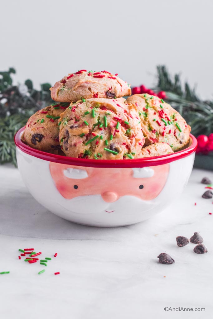 Pile of cake mix cookies in a santa design bowl with christmas decorations and candy sprinkles surrounding bowl.