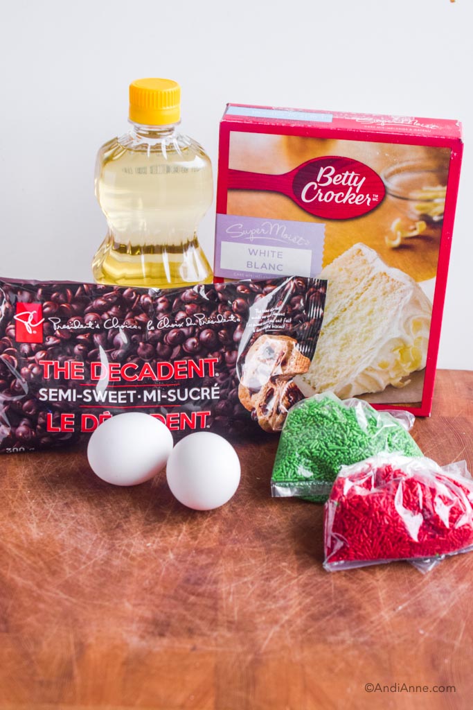 Box of white cake mix, cooking oil, chocolate chips, eggs, red and green sprinkles together on a cutting board.