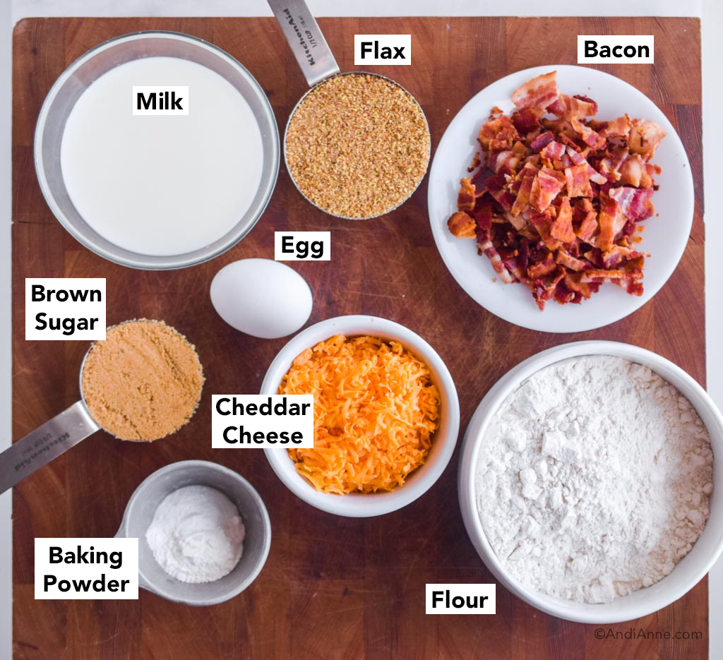 Ingredients on a cutting board: bowl of milk, cup of flax, plate of bacon, cup of brown sugar, egg, grated cheddar cheese, baking powder and flour.