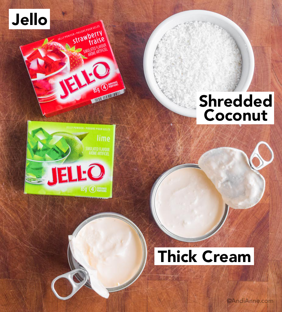 strawberry and lime jello boxes, shredded coconut in a white bowl, and two opened cans of thick cream. All sitting on a butcher block.