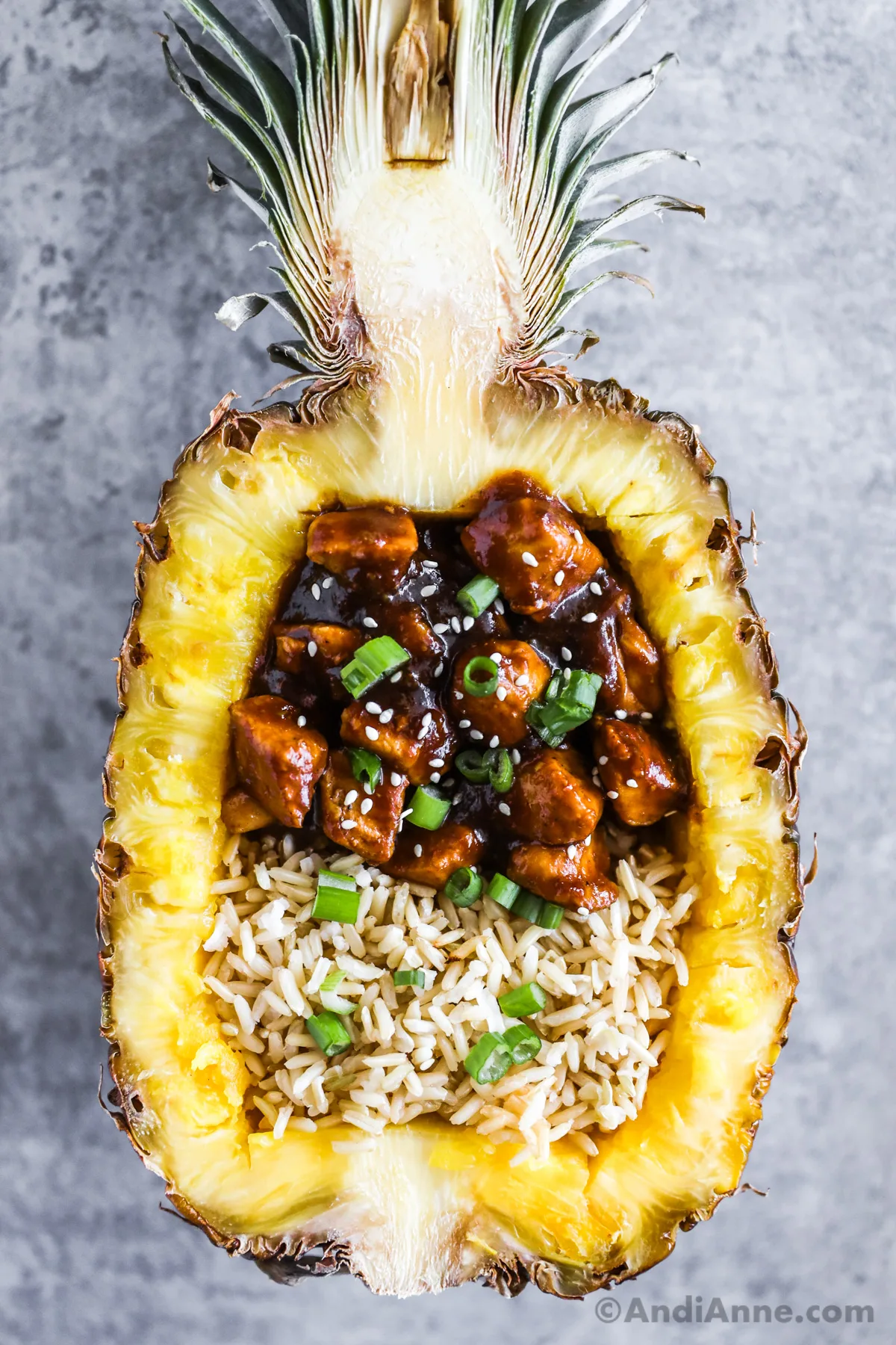 A pineapple boat with rice, teriyaki chicken bites, sprinkled with green onion and sesame seeds.