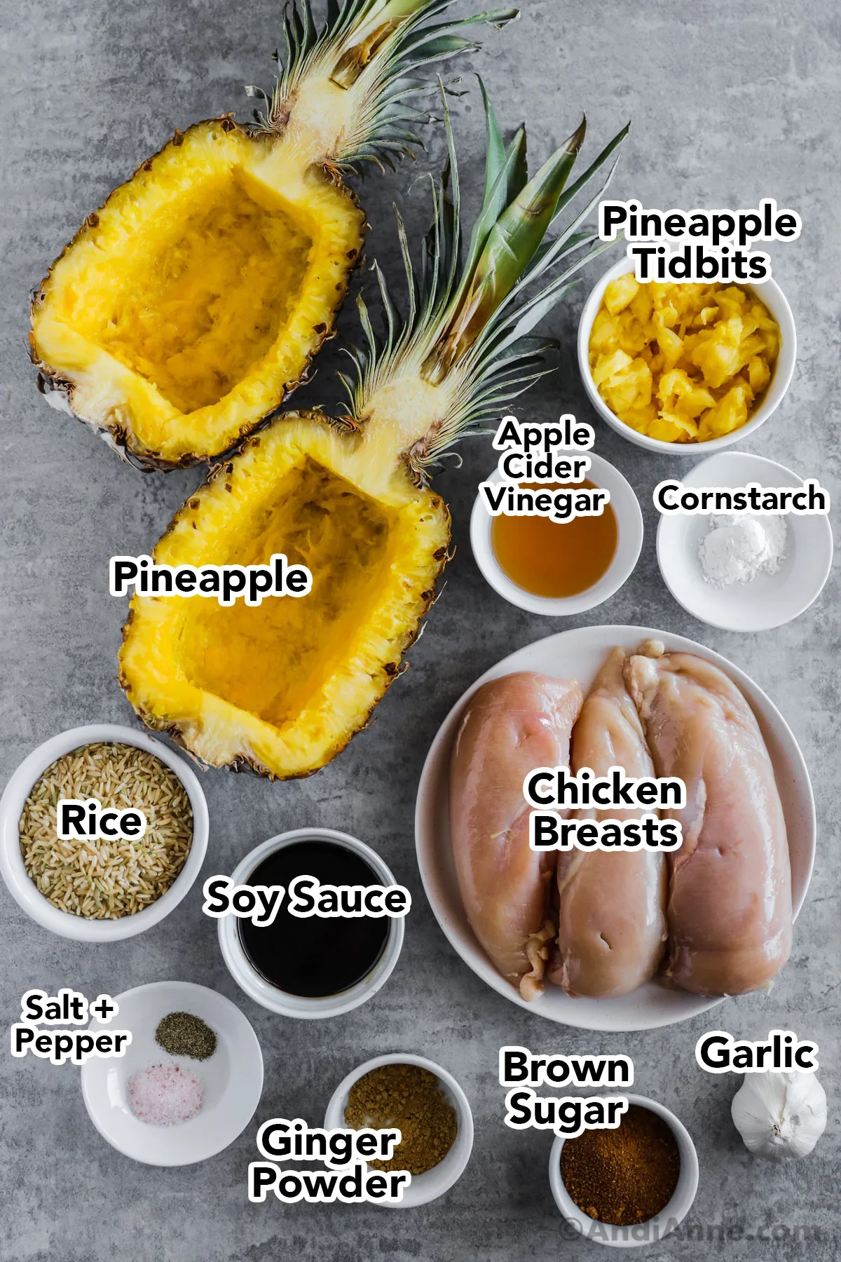 Recipe ingredients on the counter including sliced pineapple boats, bowls of apple cider vinegar, pineapple chunks, cornstarch, chicken breasts, rice, soy sauce, ginger powder, brown sugar and a garlic bulb.