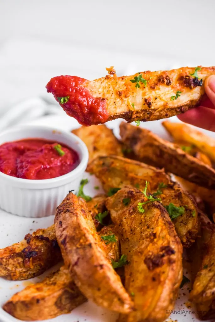 fingers holding potato wedge dipped in ketchup overtop of pile of potato wedges and ketchup in cup.