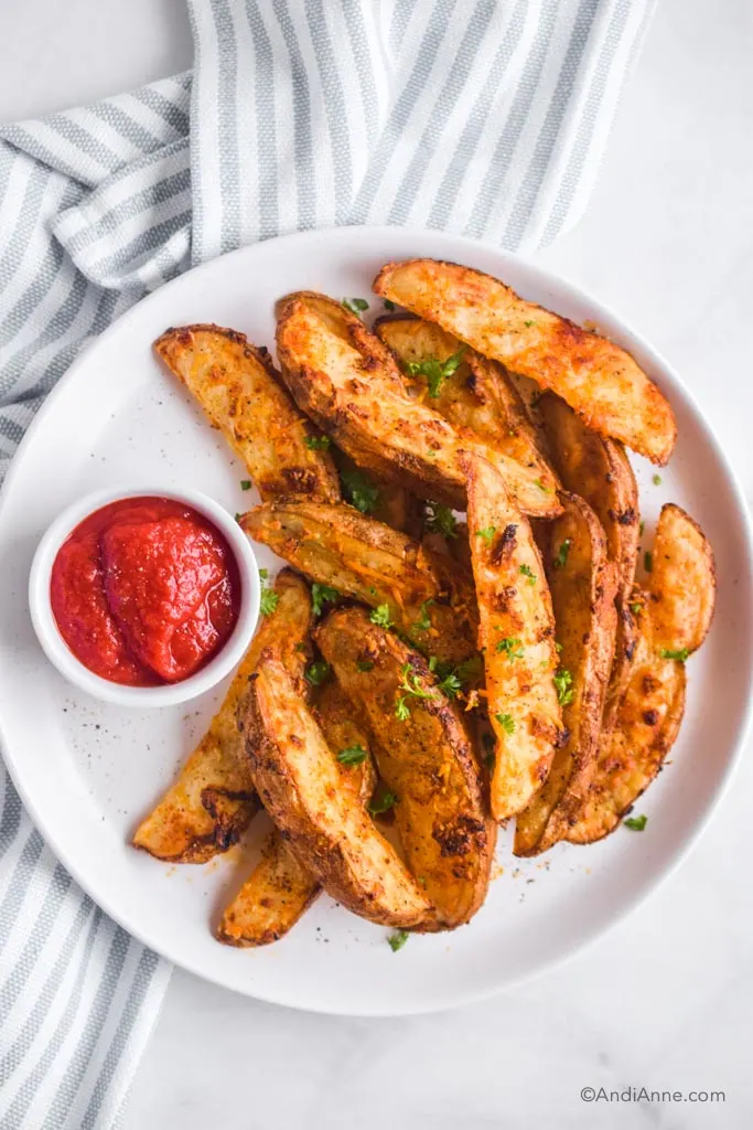crispy parmesan potato wedges with ketchup on white plate. Striped kitchen towel behind it.