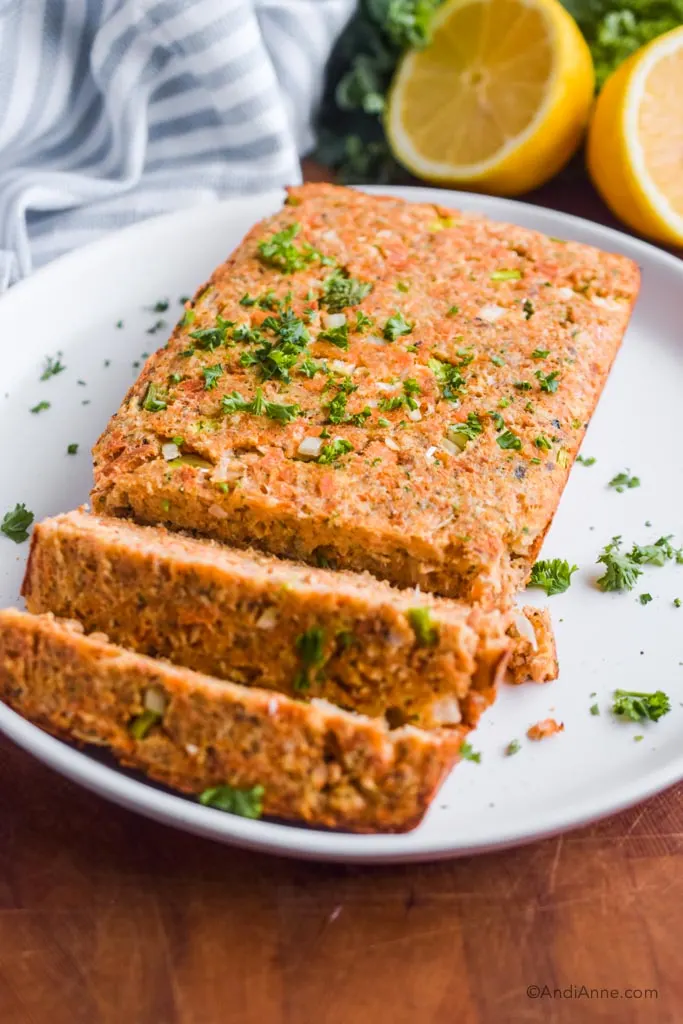 A white plate with salmon loaf with a kitchen towel, parsley and sliced lemon in the background. 