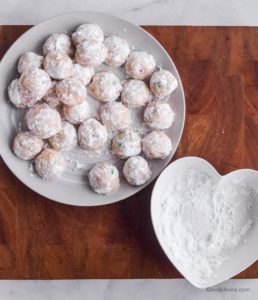 rolled cookies covered in confectioners sugar on white plate with heart plate of sugar beside it
