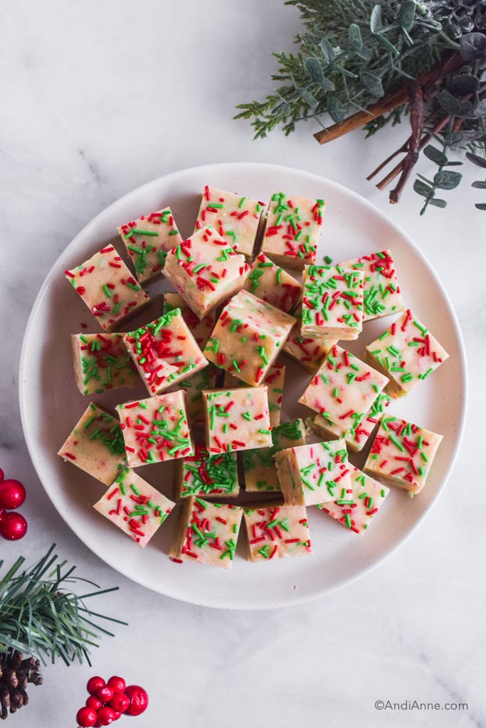 Looking down at sugar cookie fudge cut into small squares and sprinkled with red and green jimmies. Holiday greenery surrounds the sides of plate.