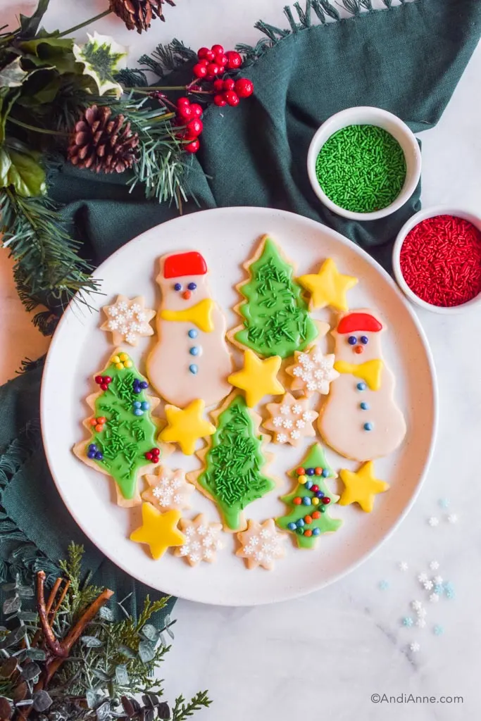 christmas sugar cookies (snowman, tree, star and snowflake shapes) on white plate with green napkin, christmas greenery and two small bowls of green and red sprinkles surrounding plate.