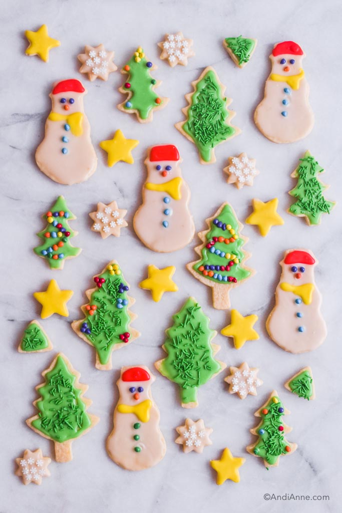 christmas sugar cookies on marble counter. Cookies include snowman shape, green trees with candy ornaments, mini snowflakes, and yellow stars.