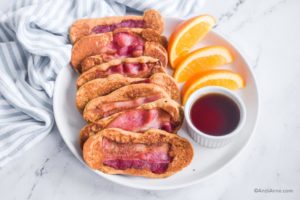 bacon pancake dippers on white plate with bowl of maple syrup and orange slices.