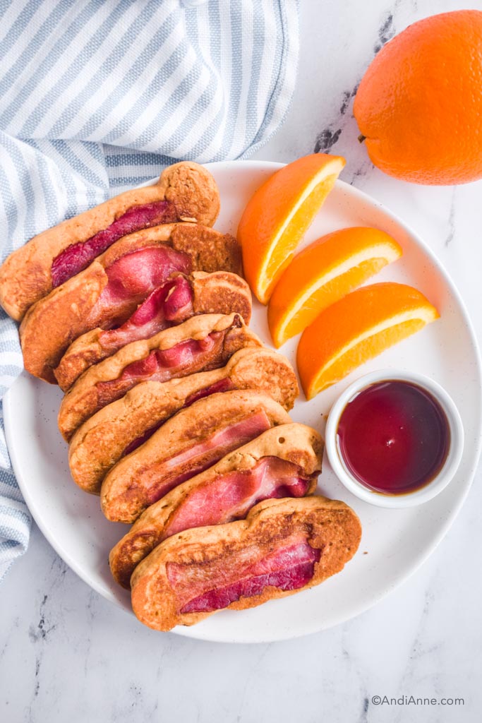 Bacon pancakes stacked on white plate with bowl of maple syrup and sliced oranges. 