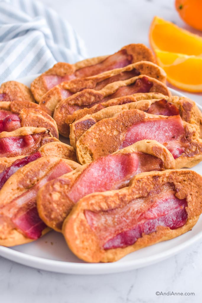 Pile of bacon pancake dippers on white plate with orange slices in background.