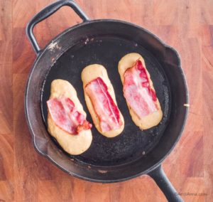 three pancake strips cooking in frying pan with strips of bacon pushed on top.