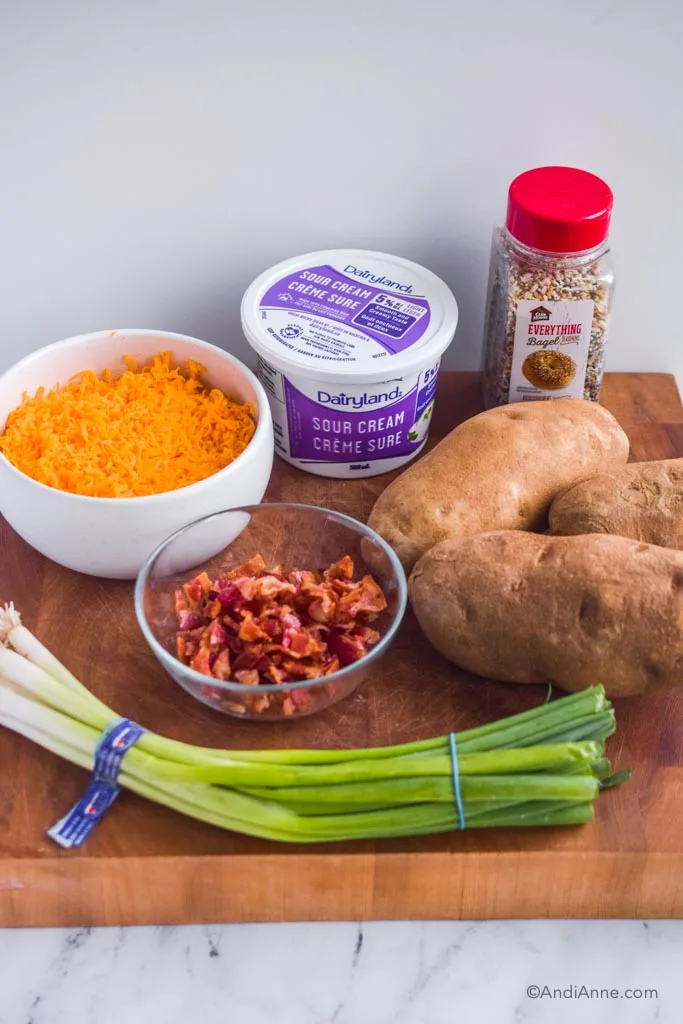 Bowl of grated cheese, container of sour cream, everything bagel seasoning, russet potatoes, bowl of crumbled bacon, and green onions.