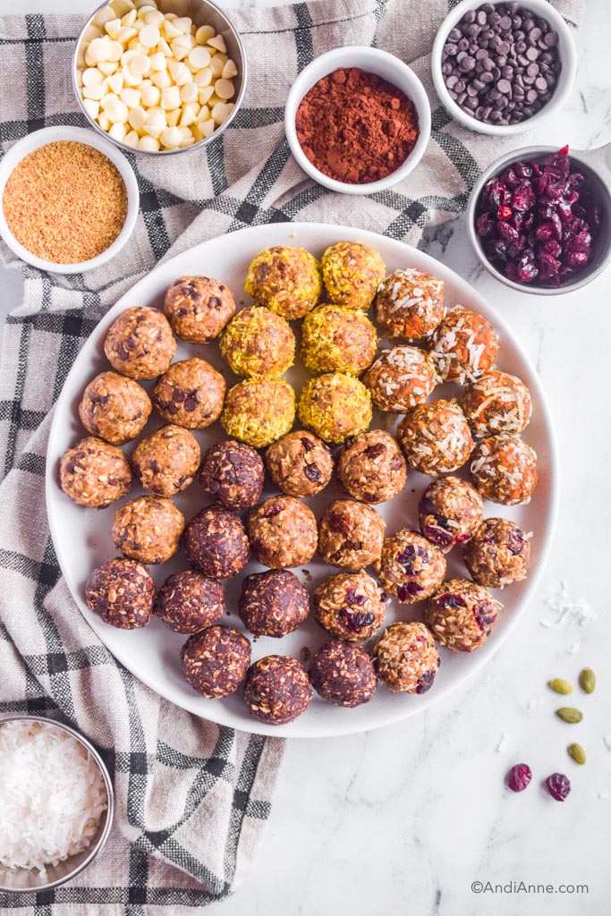 Various energy balls on white plate with small bowls of ingredients beside including ground flax, white chocolate chips, cinnamon, dried cranberries. 