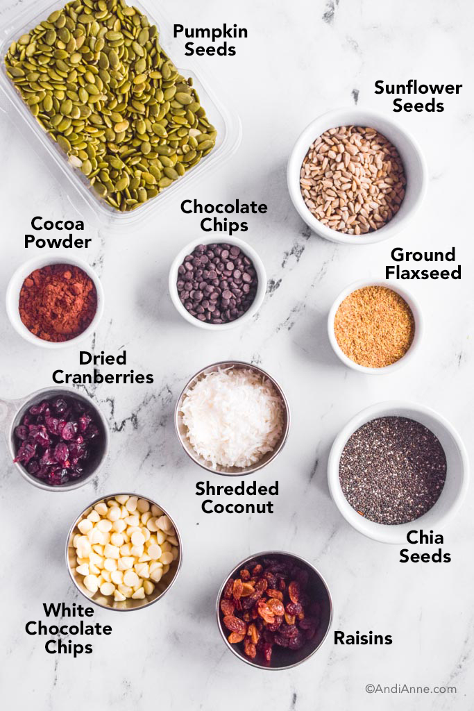 Various ingredients on white counter: container of raw pumpkin seeds, bowl of sunflower seeds, bowls of chocolate chips, cocoa powder, flaxseed, dried cranberries, shredded coconut, chia seeds, white chocolate chips, and raisins.