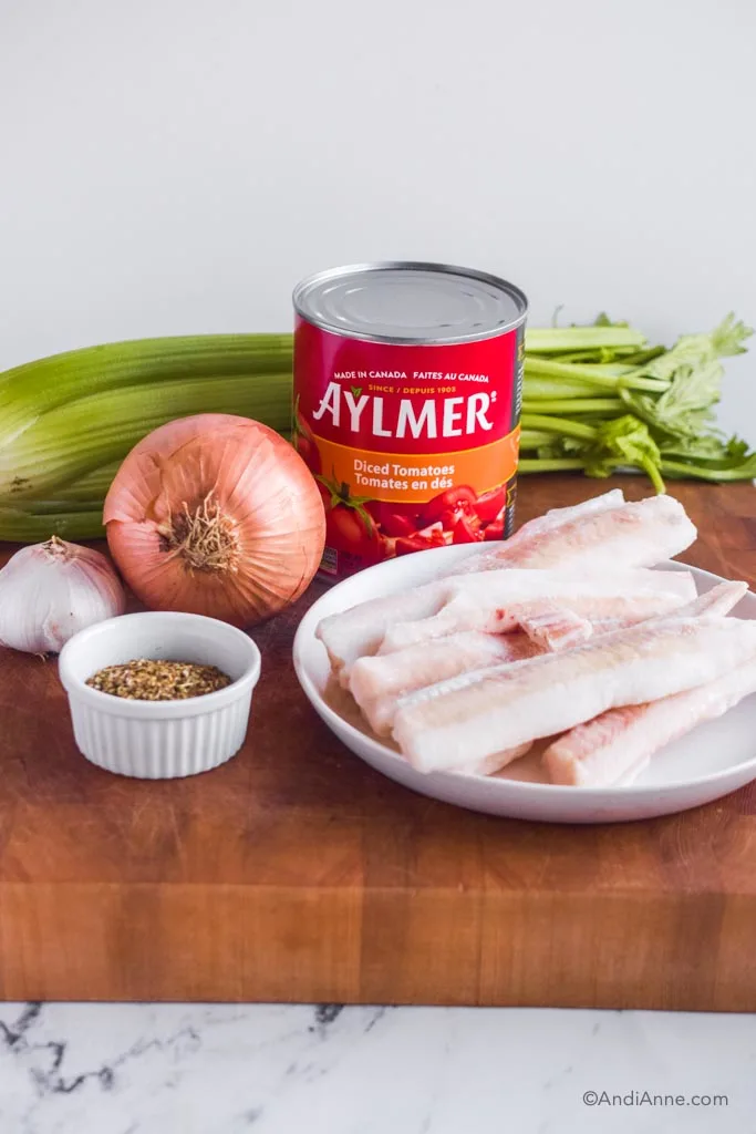 Ingredients on wood cutting board: bunch of celery, yellow onion, garlic bulb, large can diced tomatoes, small bowl of italian seasoning, plate with frozen cod fillets