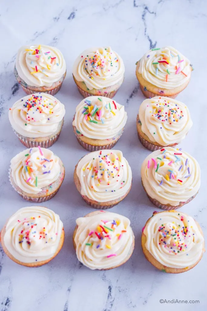 12 sprinkle cupcakes with white frosting on a marble counter.