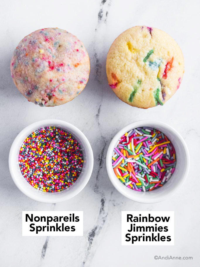 two baked cupcakes plus two bowls of rainbow sprinkles, one with nonpareils sprinkles, one with rainbow jimmies.
