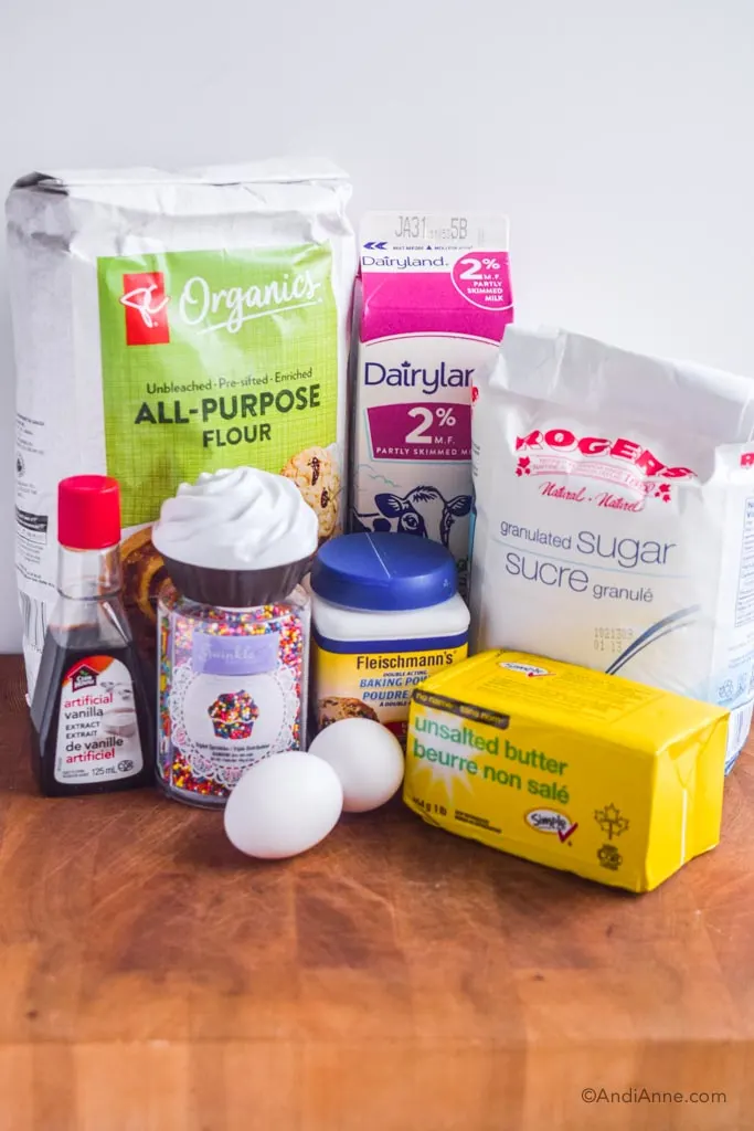 ingredients on a counter: bag of flour, carton of milk, sugar, vanilla extract, sprinkles, baking powder, unsalted butter and two eggs.