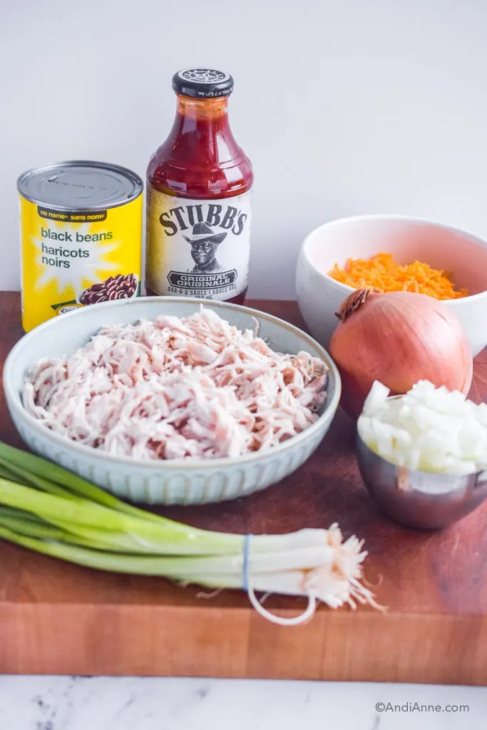 Canned black beans, jar of barbecue sauce, shredded chicken in a bowl, grated cheese, chopped yellow onion, and green onion on a wood counter.