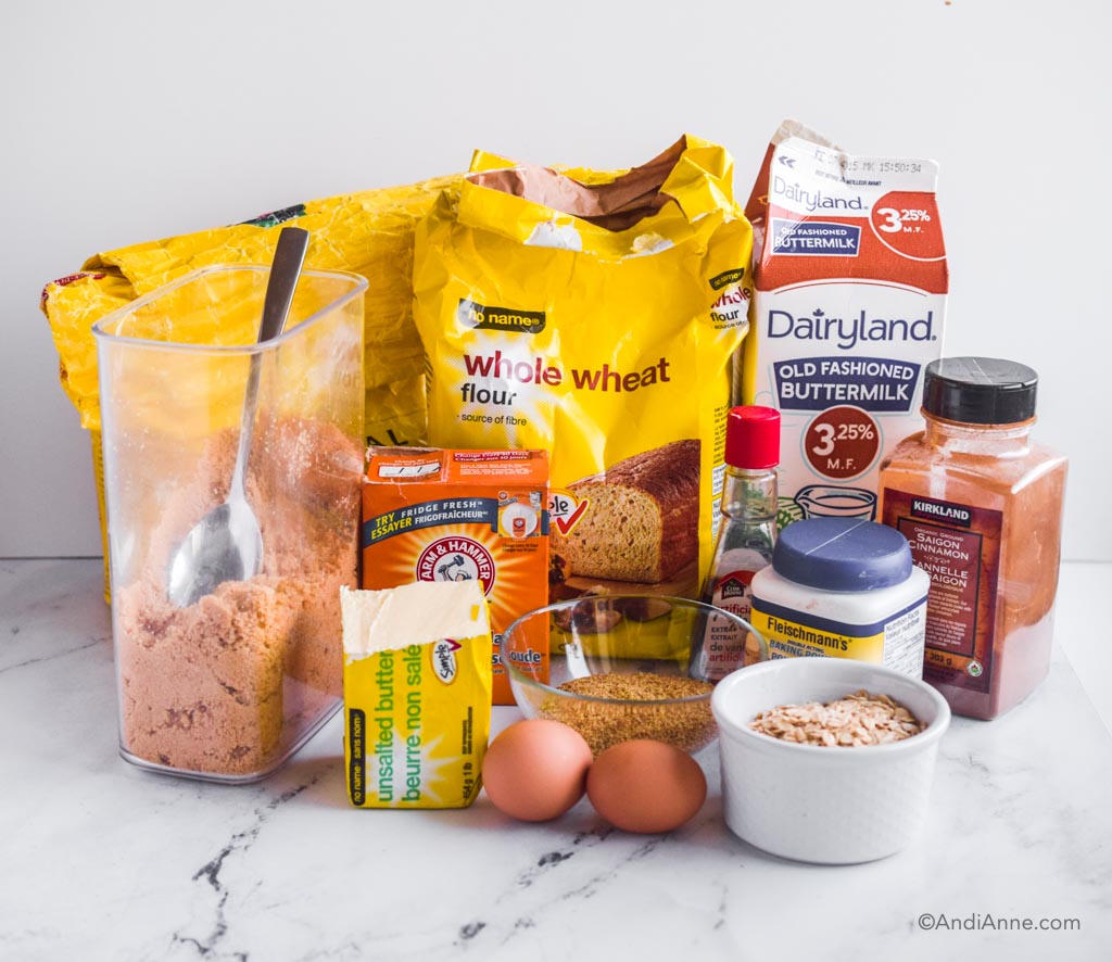 Ingredients on a white counter: wheat flour, brown sugar, baking soda, baking powder, cinnamon, vanilla extract, butter, eggs and oats.