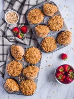 Two muffin pans with baked muffins, fresh strawberries and a kitchen towel beside.