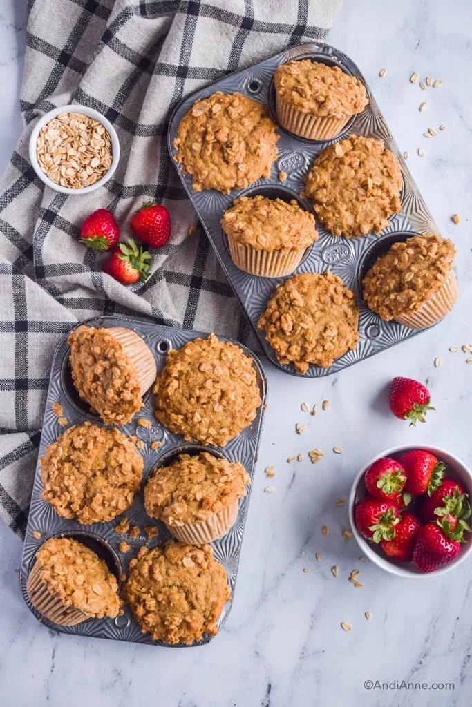 Two muffin pans with baked muffins, fresh strawberries and a kitchen towel beside.