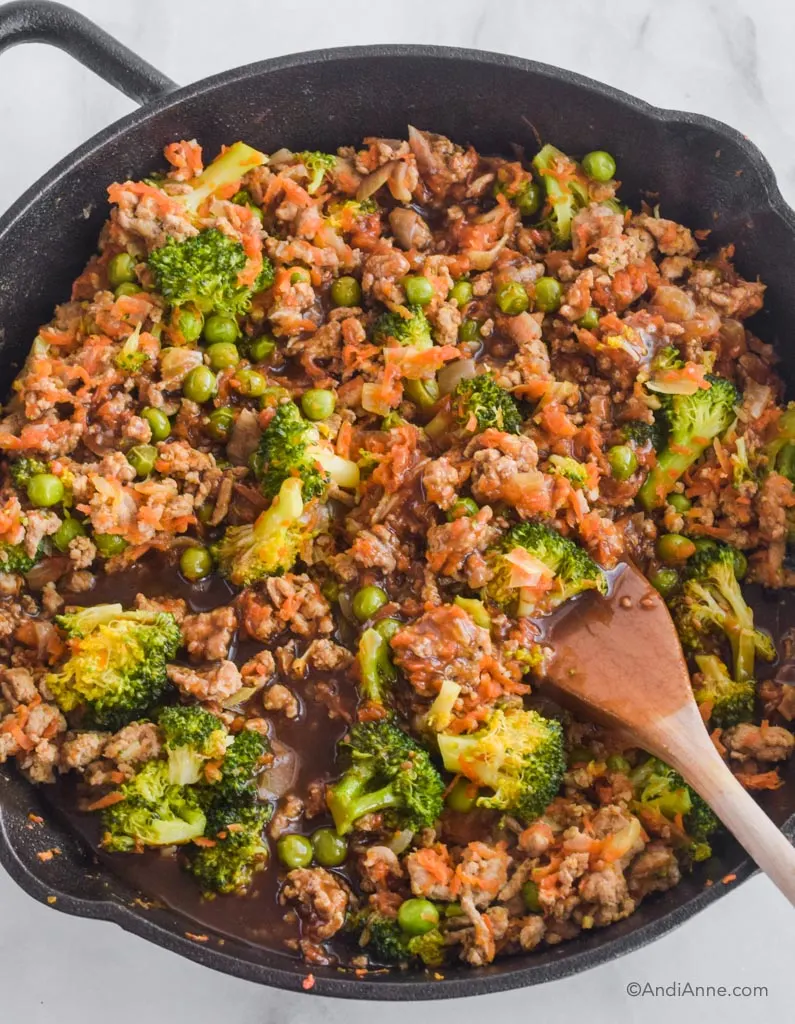 Teriyaki turkey rice bowl with broccoli, peas and shredded carrots in frying pan with wood spoon.