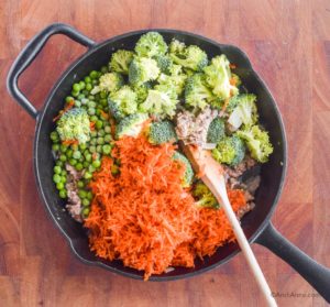 chopped broccoli, shredded carrots, peas poured on top of ground turkey in frying pan with wood spoon