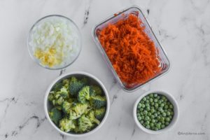 four bowls with chopped onion and garlic, shredded carrots, chopped broccoli and frozen peas