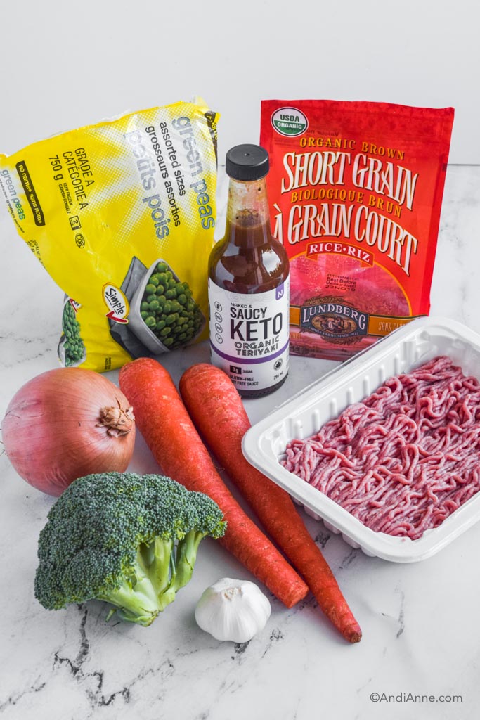 Ingredients for recipe on a table: bag of peas, keto sauce, bag of rice, onion, 2 carrots, broccoli crown, garlic bulb, and container of raw ground turkey