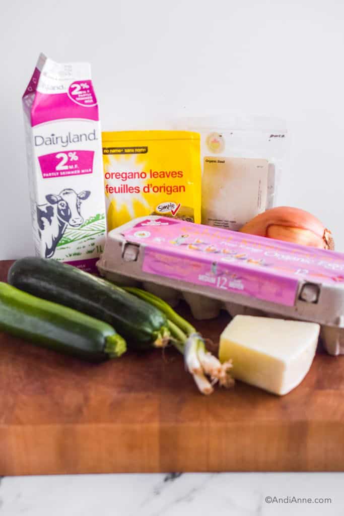 ingredients on counter for recipe: carton of milk, bake of oregano, basil, yellow onion, two zucchini, green onion, block of parmesan, carton of eggs.