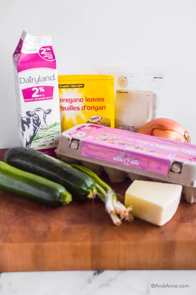 ingredients on counter for recipe: carton of milk, bake of oregano, basil, yellow onion, two zucchini, green onion, block of parmesan, carton of eggs.