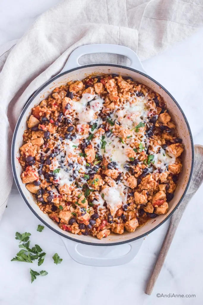Chicken burrito bowl in white pot with wood spoon, parsley and kitchen towel beside it.