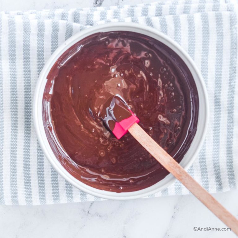 melted bakers chocolate in white bowl with mini spatula and kitchen towel underneath