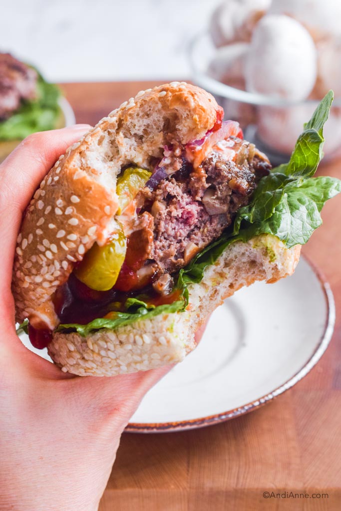 A hand holding a burger with lettuce, ketchup, pickles, onion and beef burger patty.